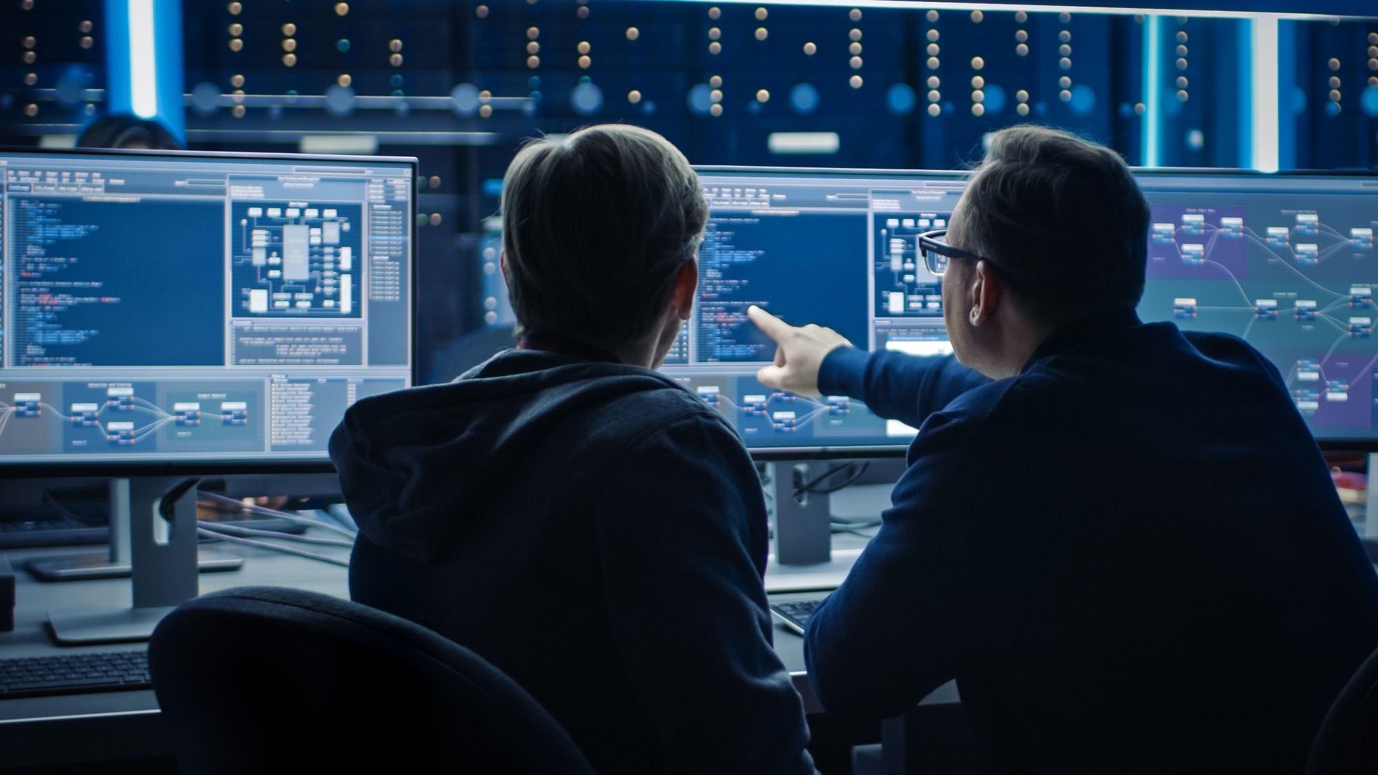 Two Professional IT Programers Discussing Blockchain Data Network Architecture Design and Development Shown on Desktop Computer Display. Working Data Center Technical Department with Server Racks Two IT experts collaborating, one pointing to a monitor, sharing security strategy as part of comprehensive data security services
