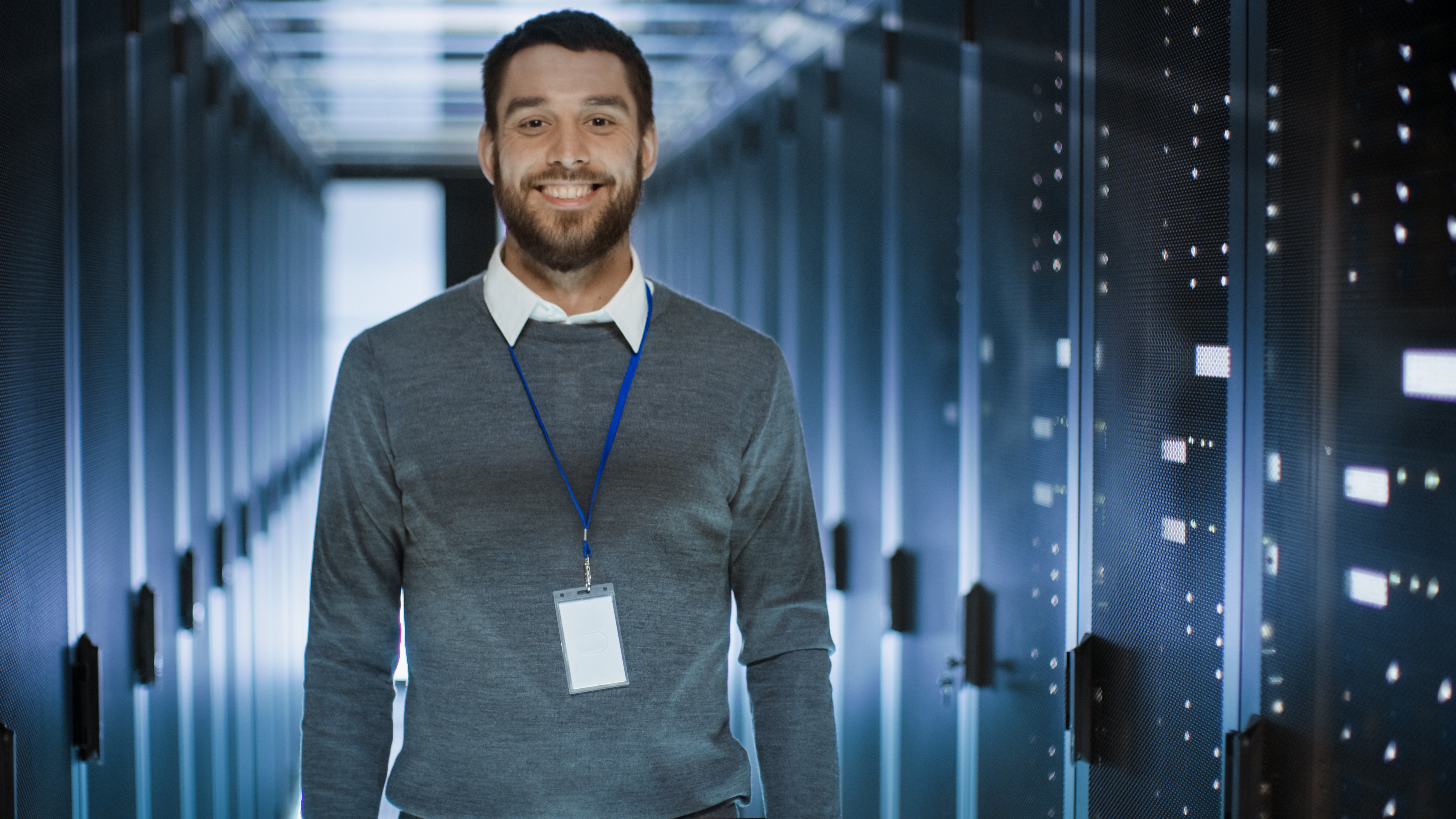 Portrait of an IT Engineer, he Smiles and Folds His Arms on a Chest. He’s Working in Data Center Full of Rack Servers. Group of IT people in a meeting concept image for IT services for financial industry